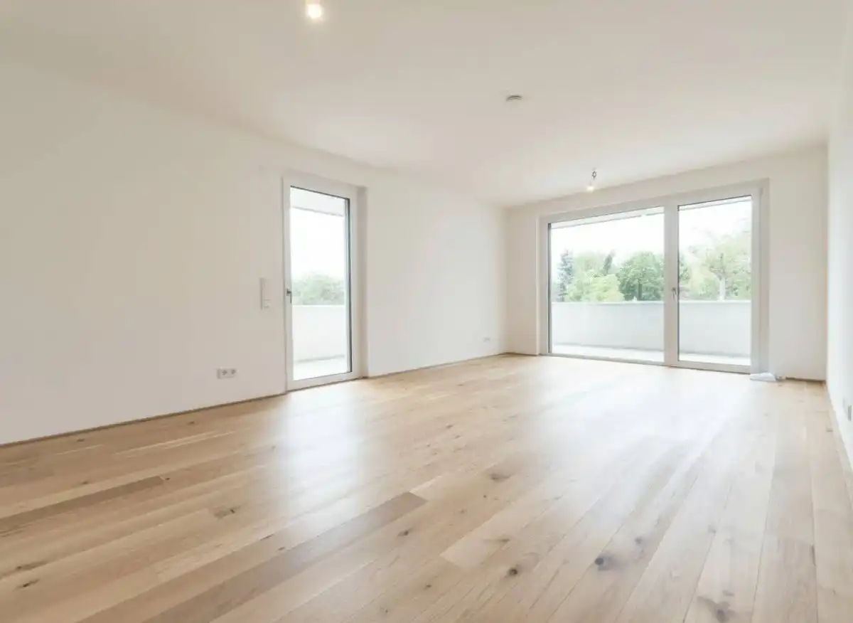 Geräumiges, unmöbliertes Zimmer mit hellem Holzboden und großen Fenstern zum Balkon.