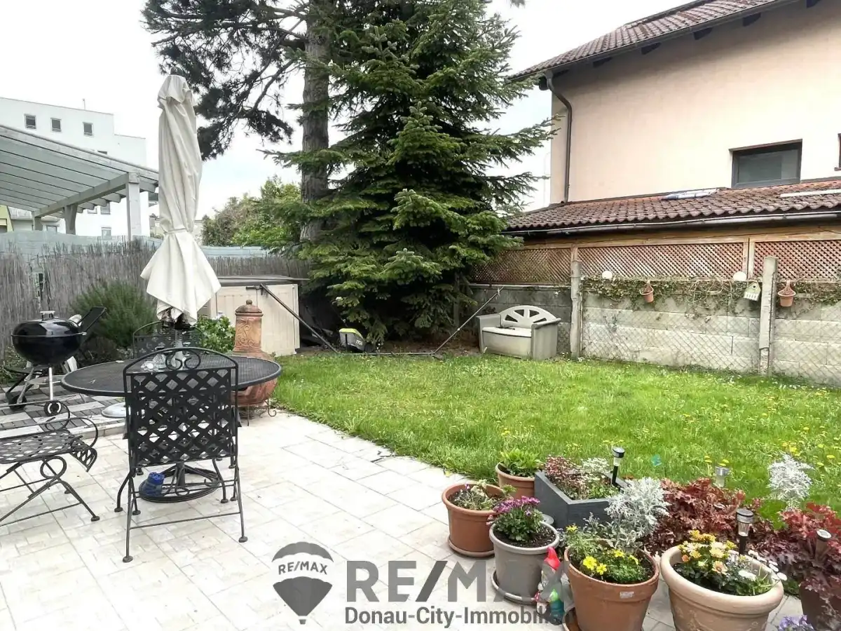 Gepflegter Gartenbereich mit einer Terrasse, Sitzgelegenheiten und einem großen Baum.