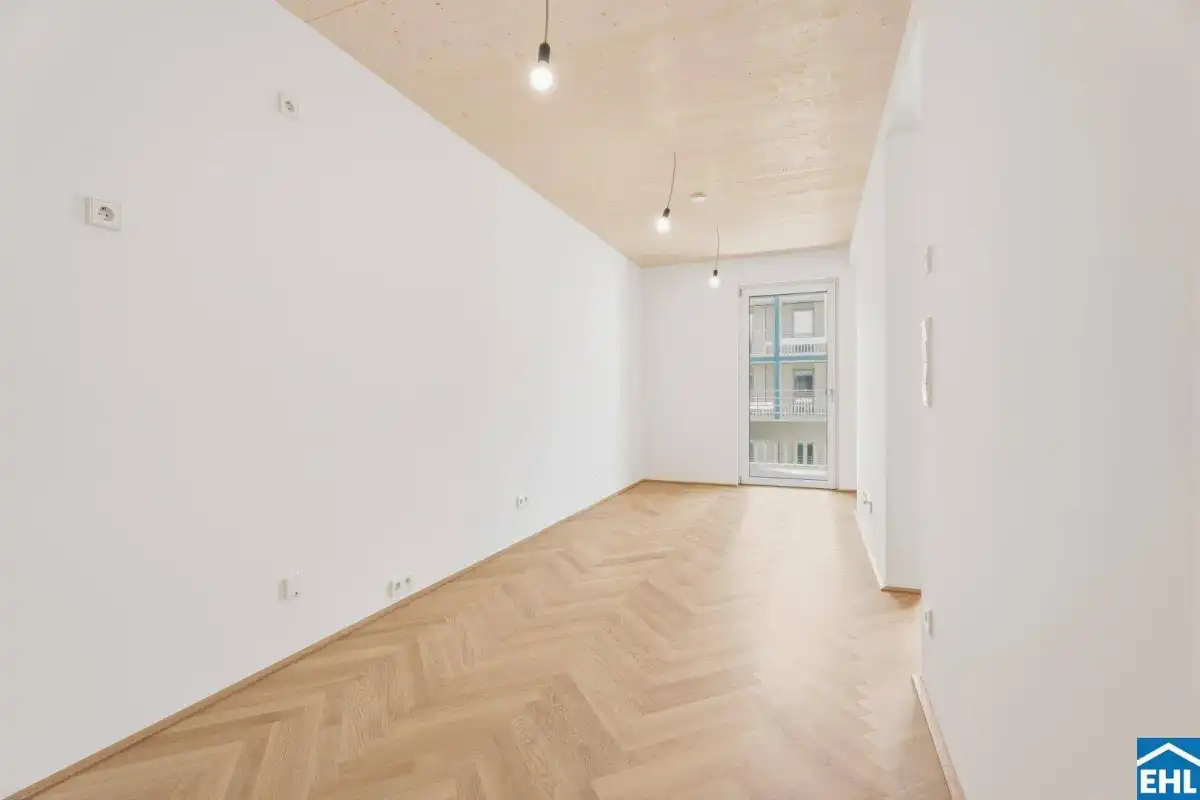 Lichtdurchfluteter Raum mit Fischgrätparkett, Holzbalkendecke und Zugang zu einem Balkon.