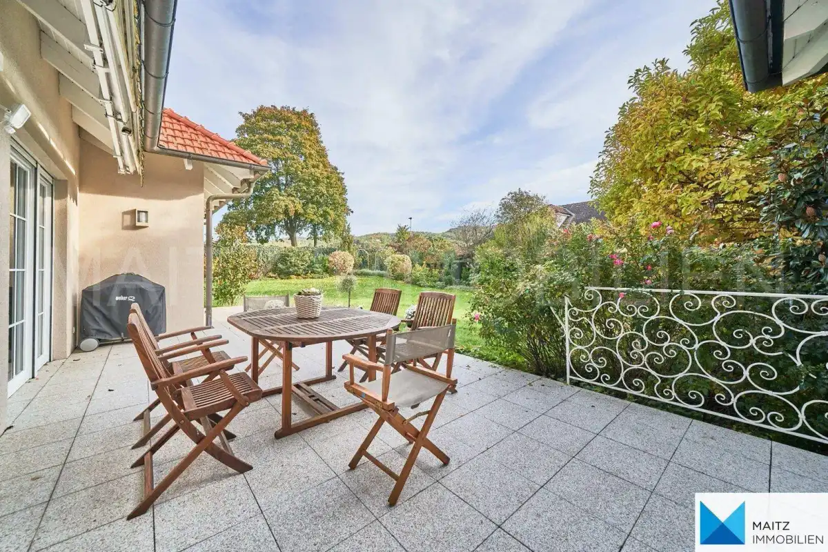 Geräumige Terrasse mit Holztisch und Stühlen, umgeben von üppigem Grün.