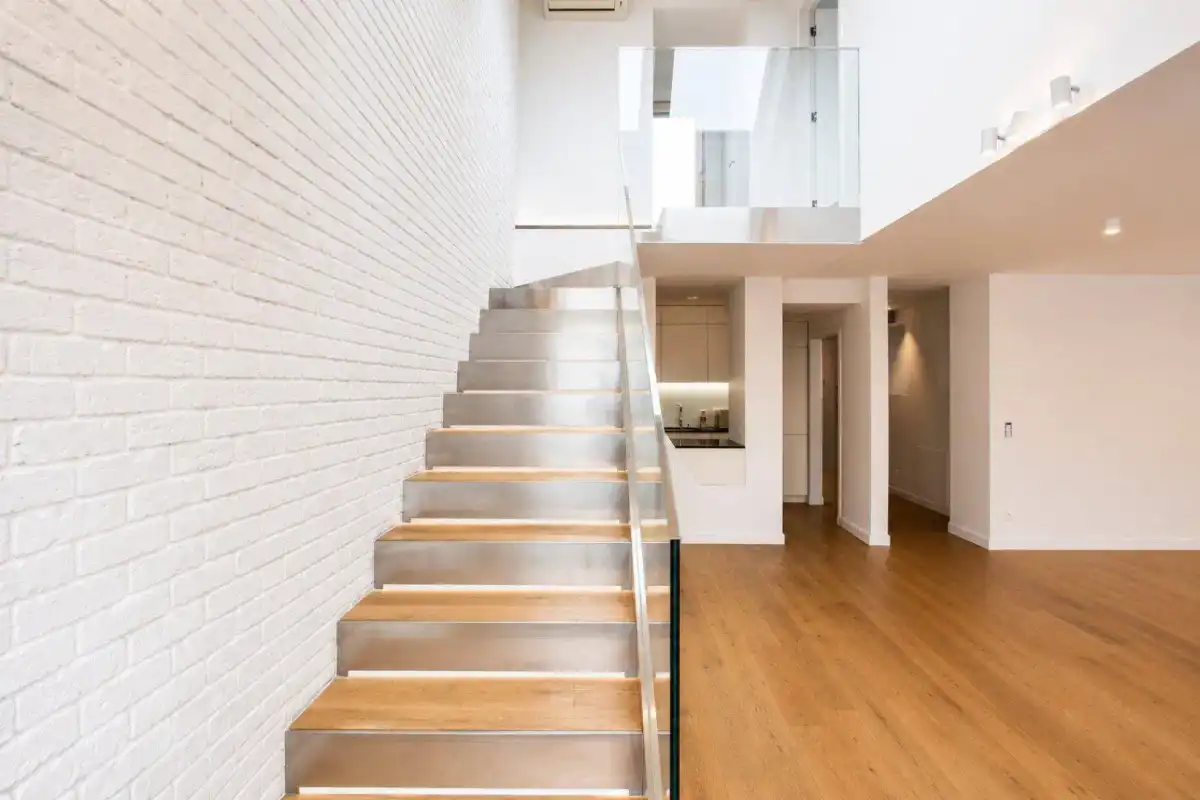 Moderne Treppe mit Holzstufen und Glasgeländer in einem hellen Innenbereich mit weißer Ziegelwand.