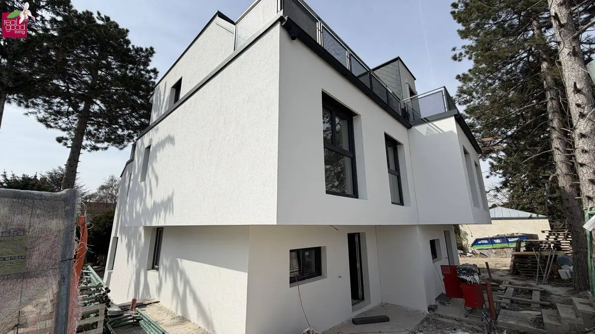 Moderne weiße Hausfassade eines Neubaus mit großen Fenstern, umgeben von Bäumen.