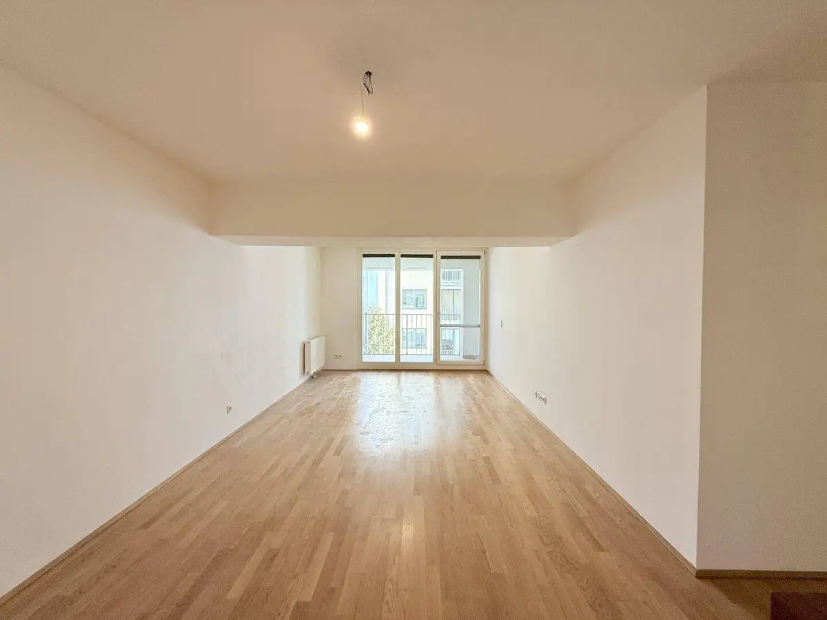 Helles, leeres Wohnzimmer mit Parkettboden und Zugang zu einem Balkon oder einer Loggia.