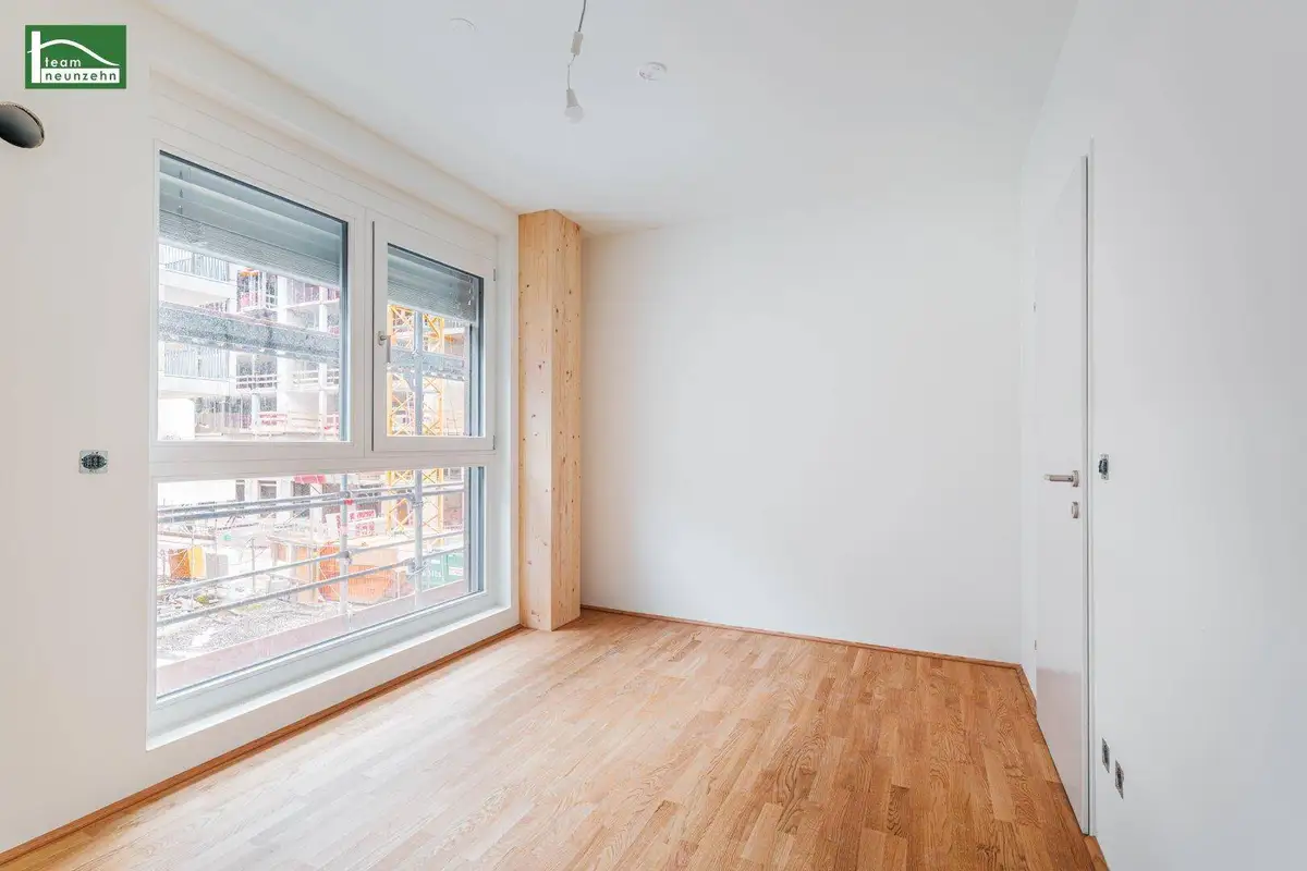 Heller, leerer Raum mit Holzboden und großem Fenster, das auf eine Baustelle blickt.