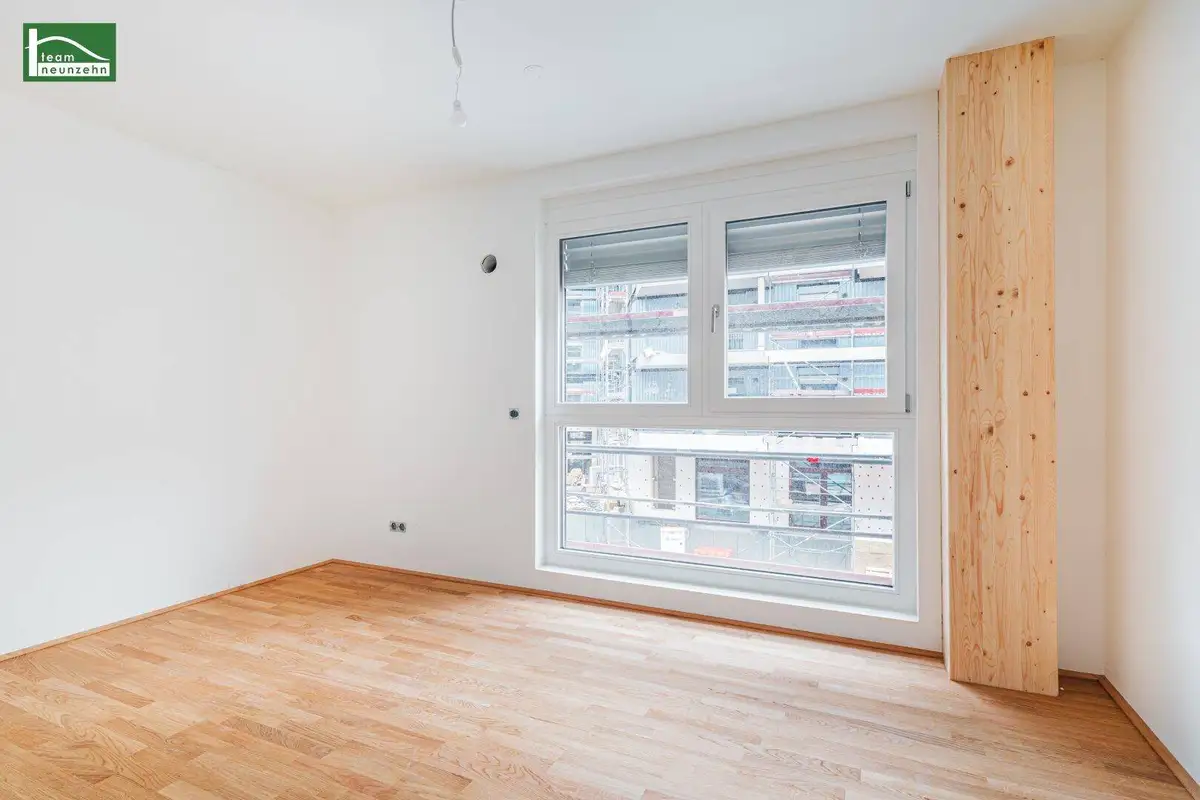 Leerer Raum mit hellem Holzboden und Fenster, das auf eine Baustelle blickt.