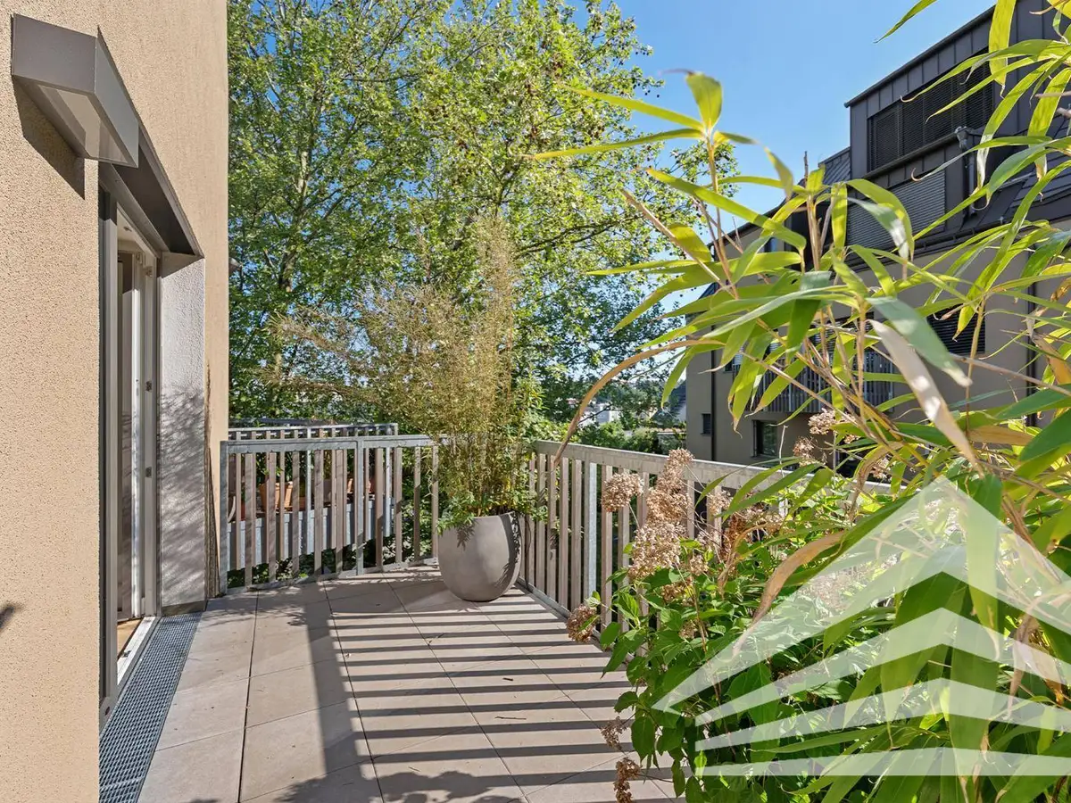 Sonniger Balkon mit Fliesenboden, Pflanzen und Blick auf grüne Bäume und umliegende Gebäude.